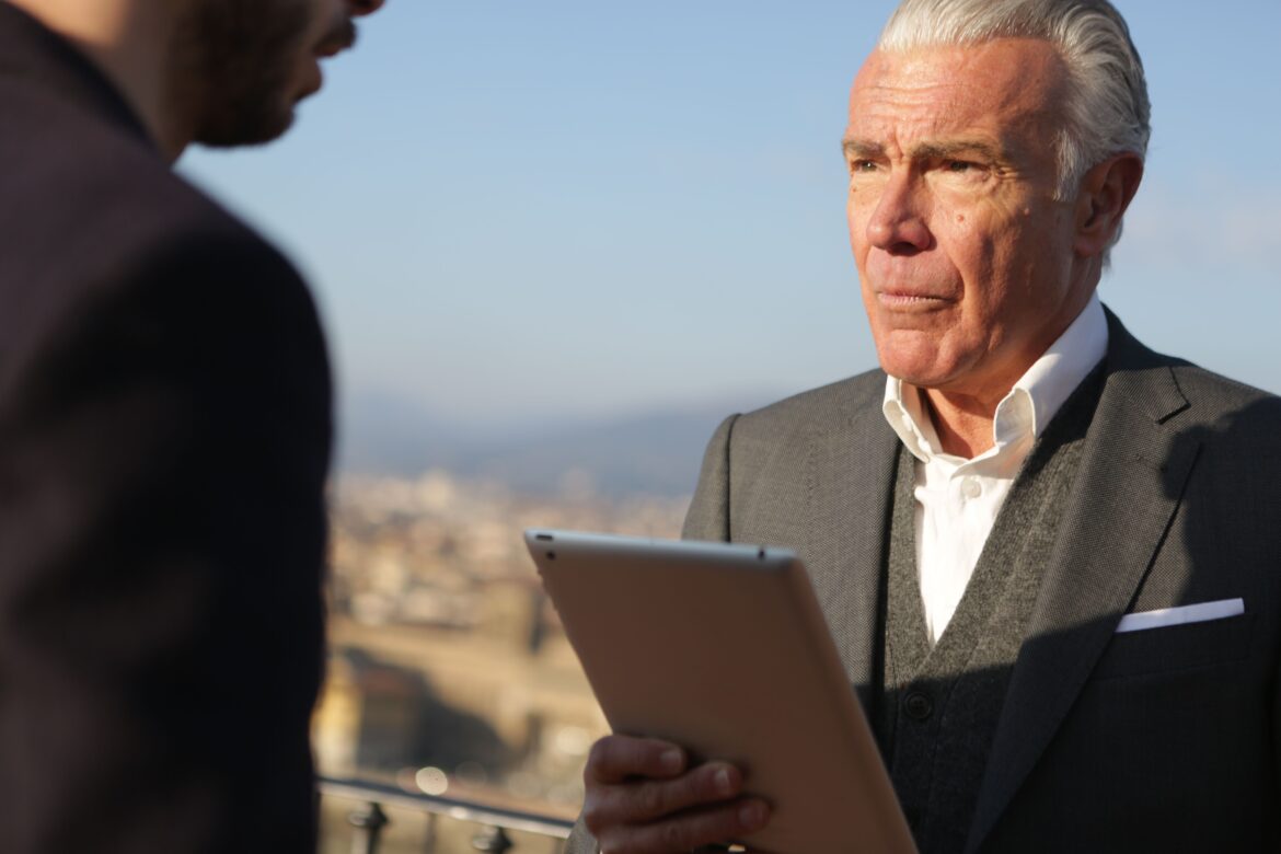Man in black suit holding silver ipad looking at another man