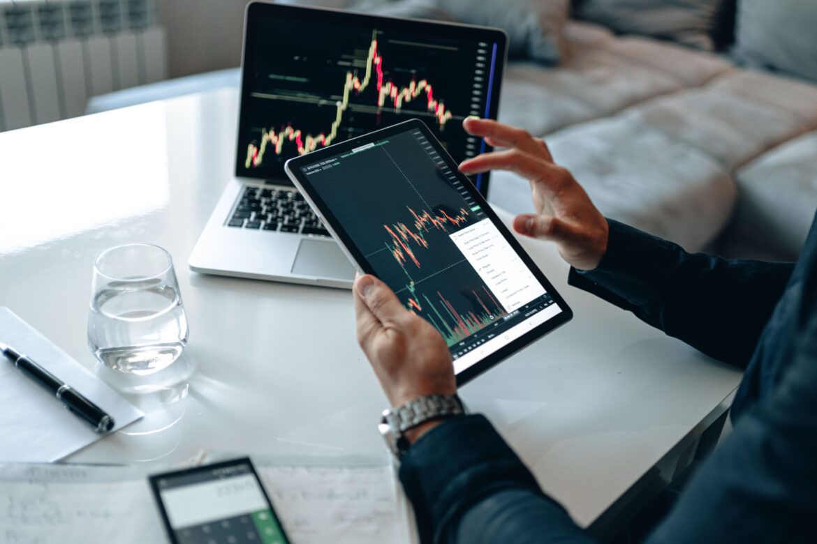 A person in blue long sleeves holding a tablet with cryptocurrency graph neat laptop on the table
